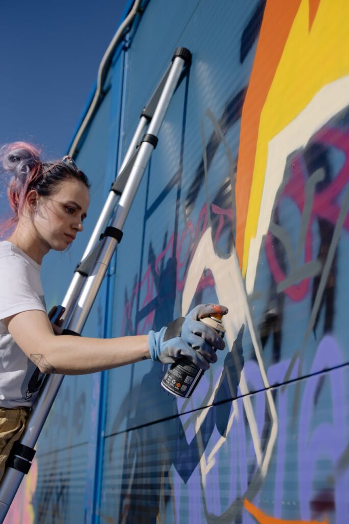 concentrated young lady using spray can while painting graffiti wall on street in sunlight